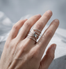 A men's architectural silver ring featuring an intricate interplay of lines inspired by the structure of a dragonfly's wing. The piece stands upright on a rough, textured gray concrete pedestal. Directed studio lighting highlights the raw beauty and mirror-like polish of the metal, creating sharp, geometric shadows. In the background are soft, blurred white translucent accents, symbolizing the element of ether and pristine nature.