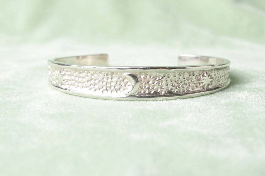 Minimalist silver cuff bracelet with moon and star symbols isolated on a soft light green background, showcasing smooth edges and premium polishing.
